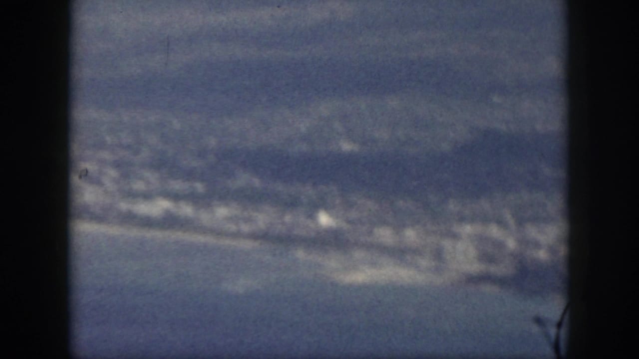 Oahu coastline in a soft 1959 8mm aerial view, with ocean and town detail blurred by motion and haze.