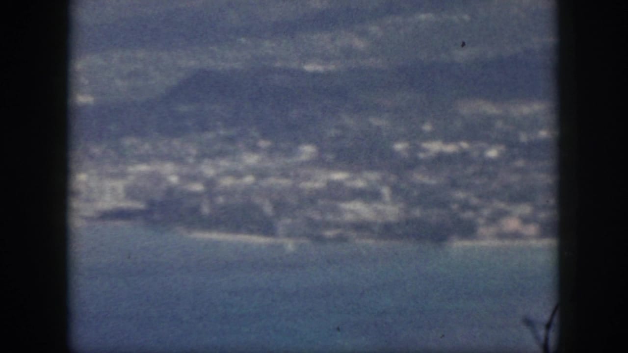 1959 Oahu aerial home-movie frame showing shoreline, low hills, clustered buildings, and blue water.