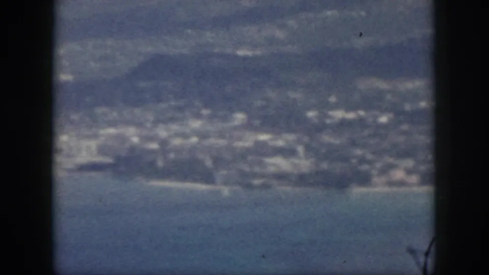 1959 Oahu aerial home-movie frame showing shoreline, low hills, clustered buildings, and blue water.