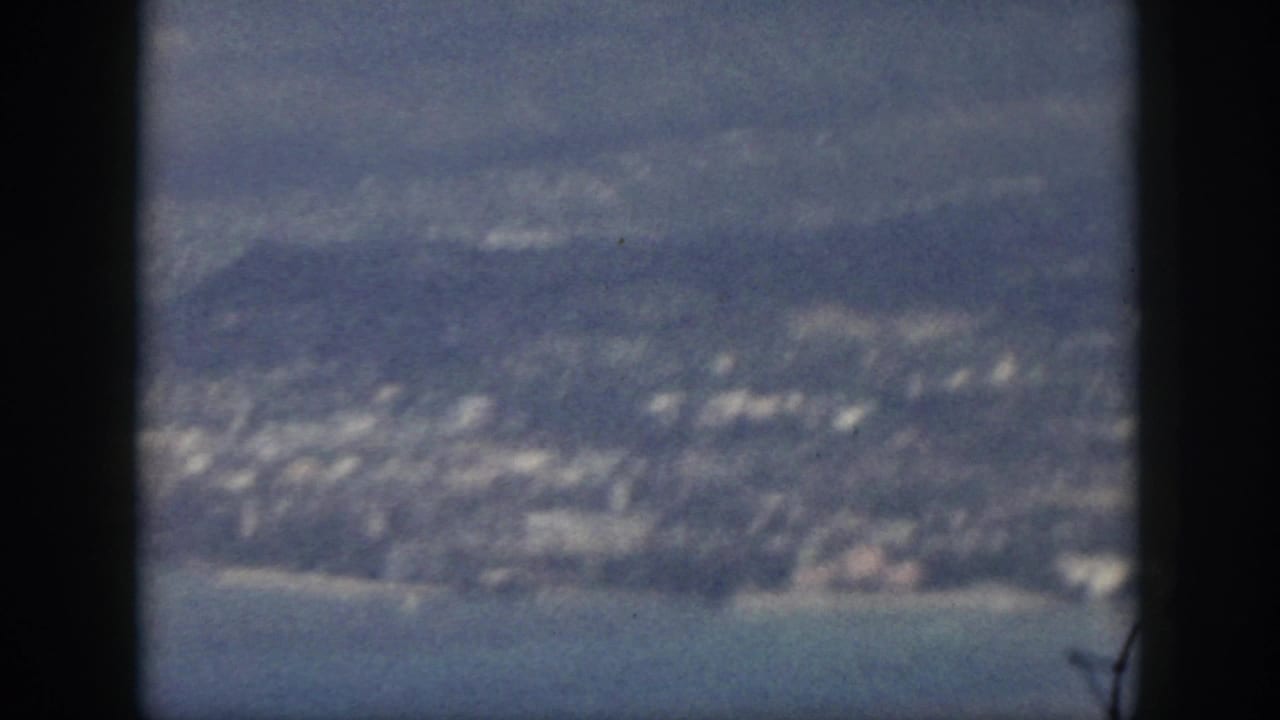 1959 Oahu coastal settlement and shoreline seen from above, with mountains in haze and a dark vertical band crossing the frame.