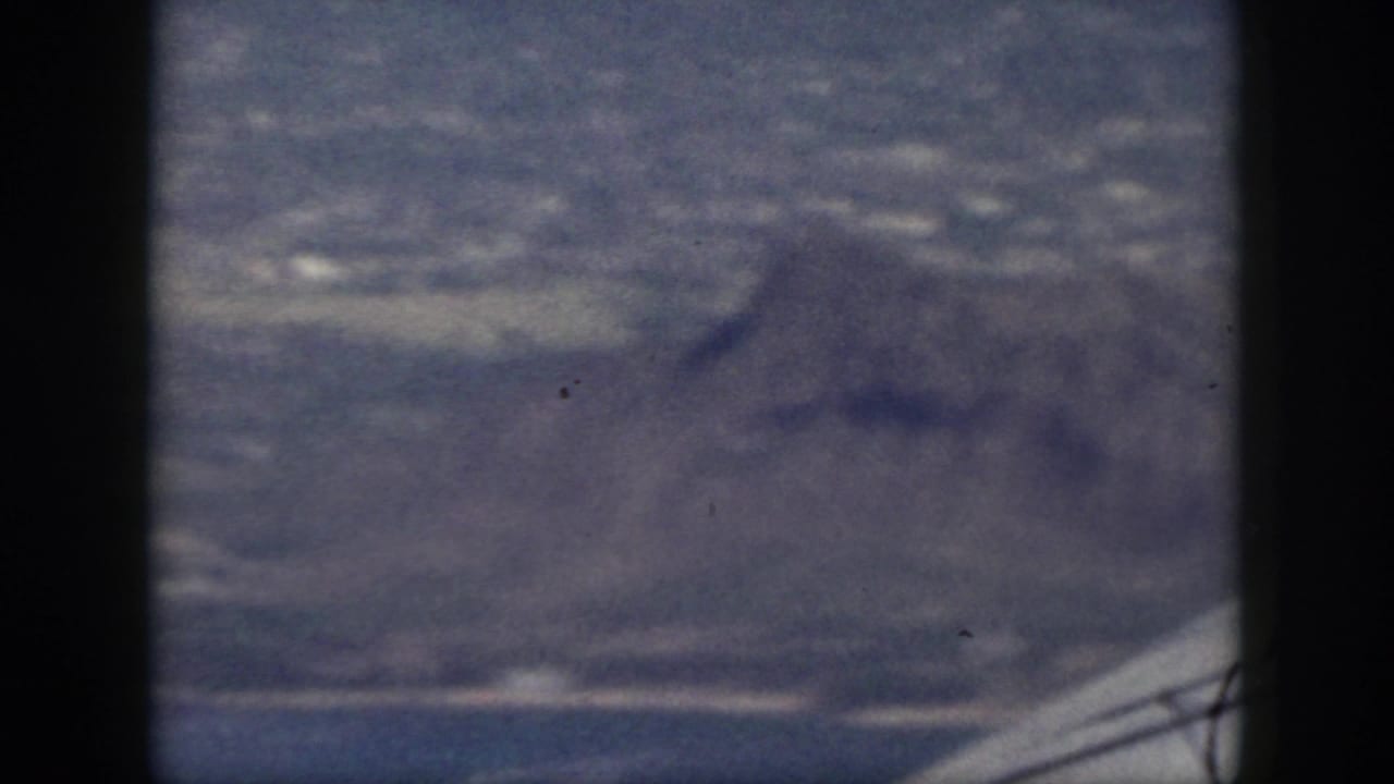 Blue-tinted 8mm view of Oahu terrain, with a mountain slope in the foreground and coastal development beyond.