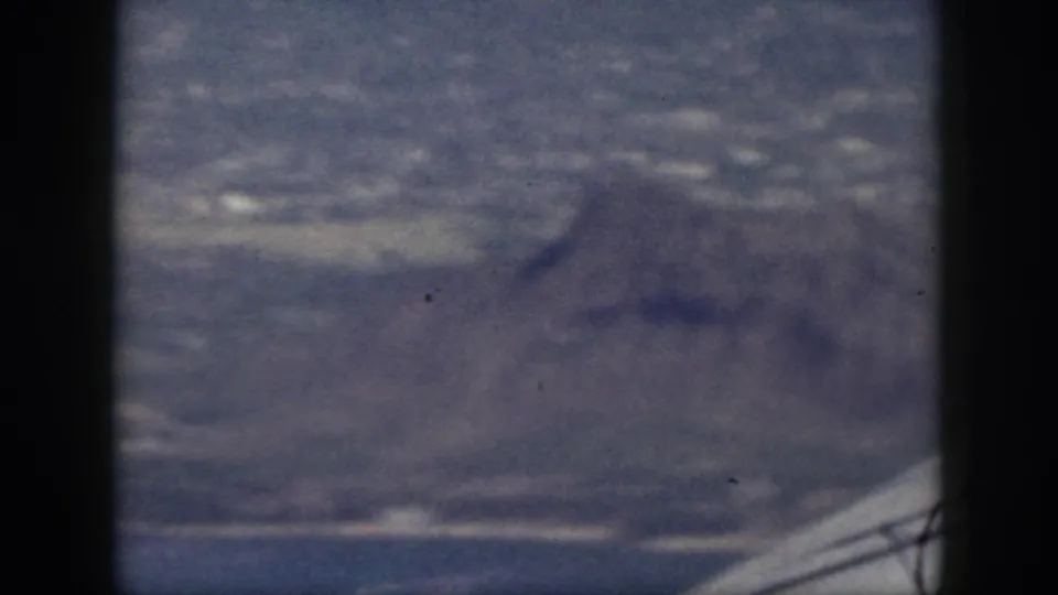 Blue-tinted 8mm view of Oahu terrain, with a mountain slope in the foreground and coastal development beyond.