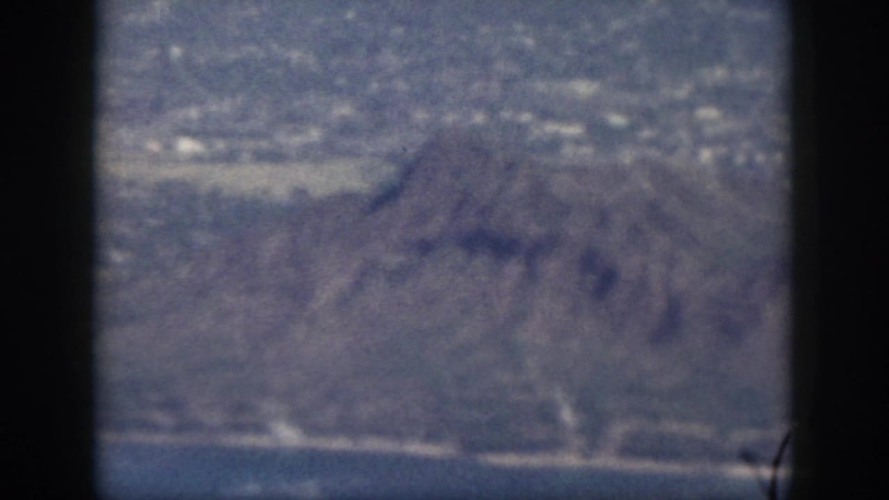 1959 Oahu aerial frame showing rugged mountain terrain below, with coastal town structures and water in the distance.