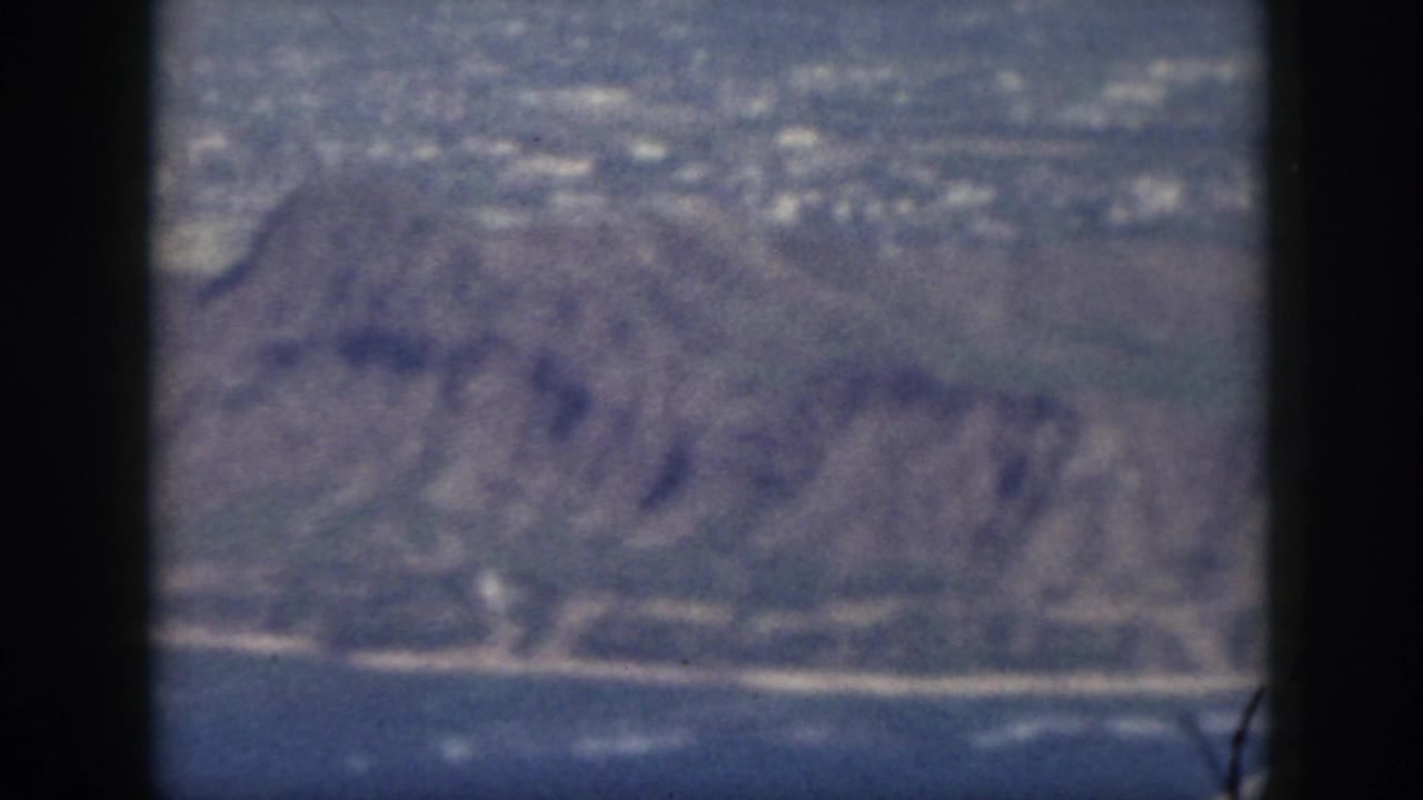 Soft aerial view of rugged coastline, hills, ocean, and dark aircraft window dividers.