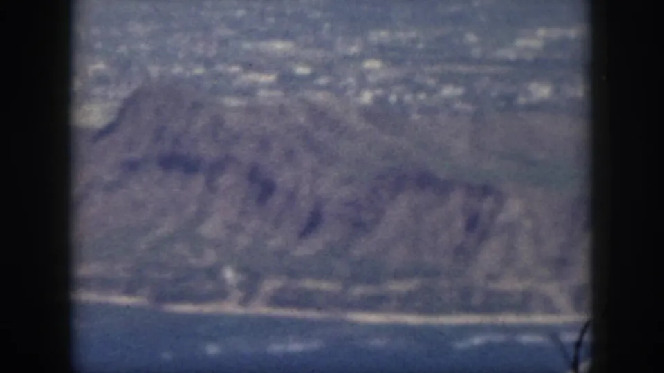 1959 Oahu aerial 8mm frame showing a blurred rocky shoreline, hillside terrain, ocean edge, and aircraft window framing.
