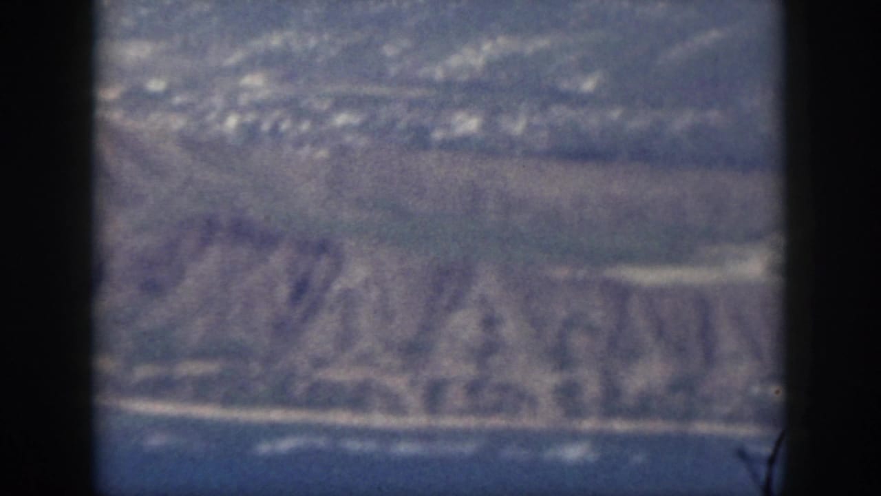 Aerial shoreline view with hills, ocean edge, scattered structures, and dark window framing.