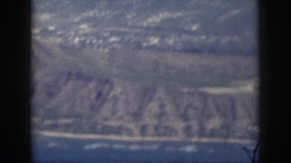 1959 Oahu aerial frame looking through aircraft windows at a mountainous shoreline with scattered settlement near the coast.