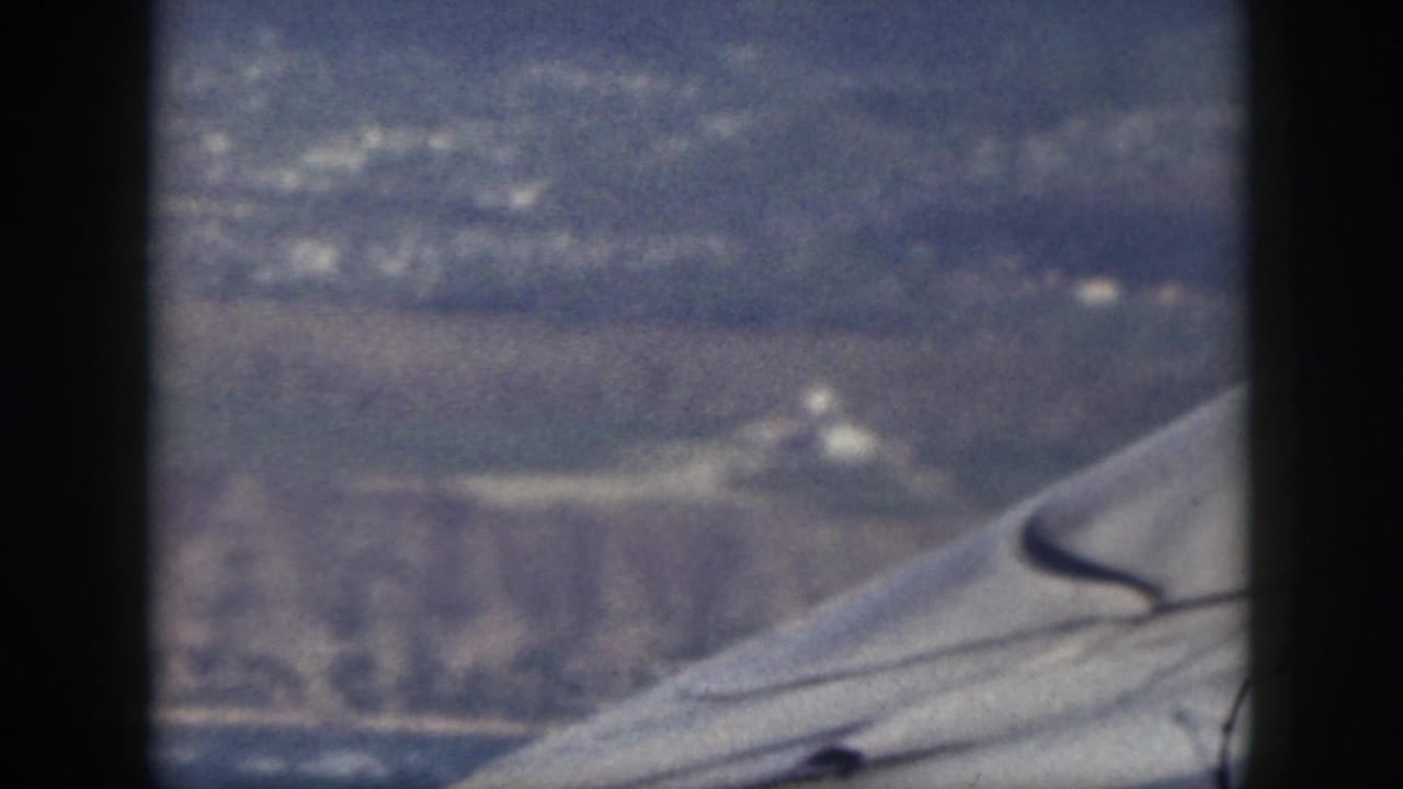 1959 Oahu aerial 8mm frame with an aircraft wing crossing the foreground above coastal hills, open land, and blue water.