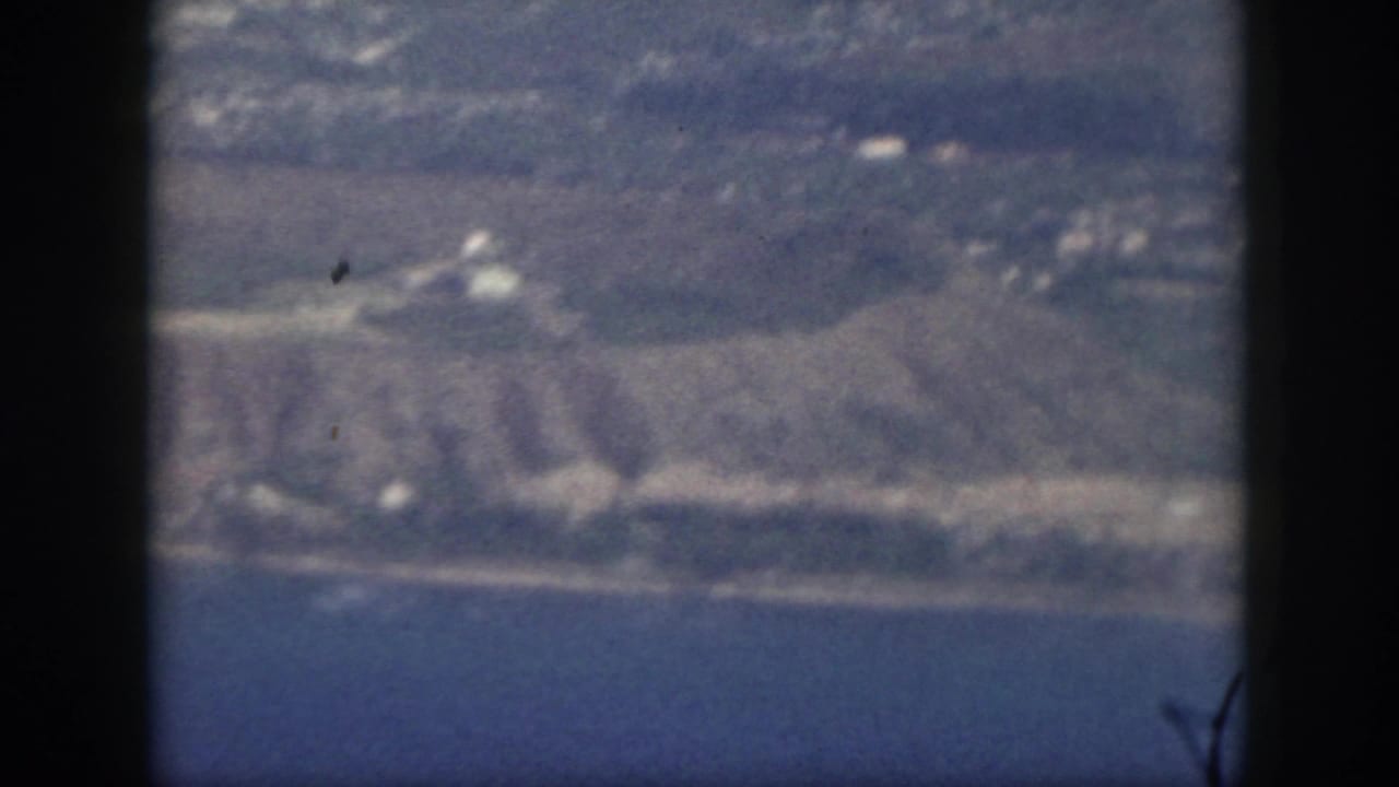 1959 Oahu aerial frame showing blue water, a pale coast, low hills, open land, and small shoreline structures through aircraft framing.