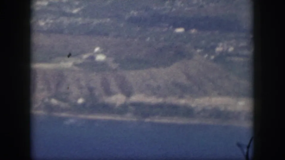 1959 Oahu aerial frame showing blue water, a pale coast, low hills, open land, and small shoreline structures through aircraft framing.