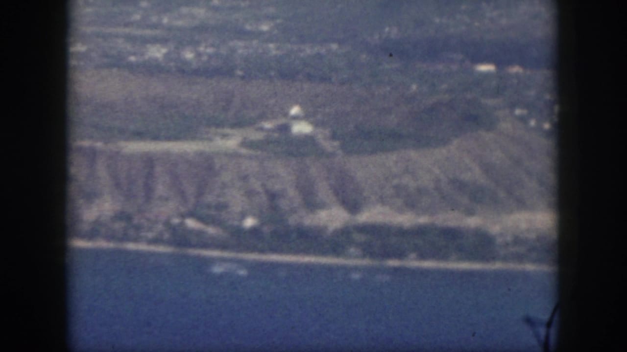 1959 Oahu aerial home-movie frame showing coastline, water, and distant land through dark airplane window dividers.