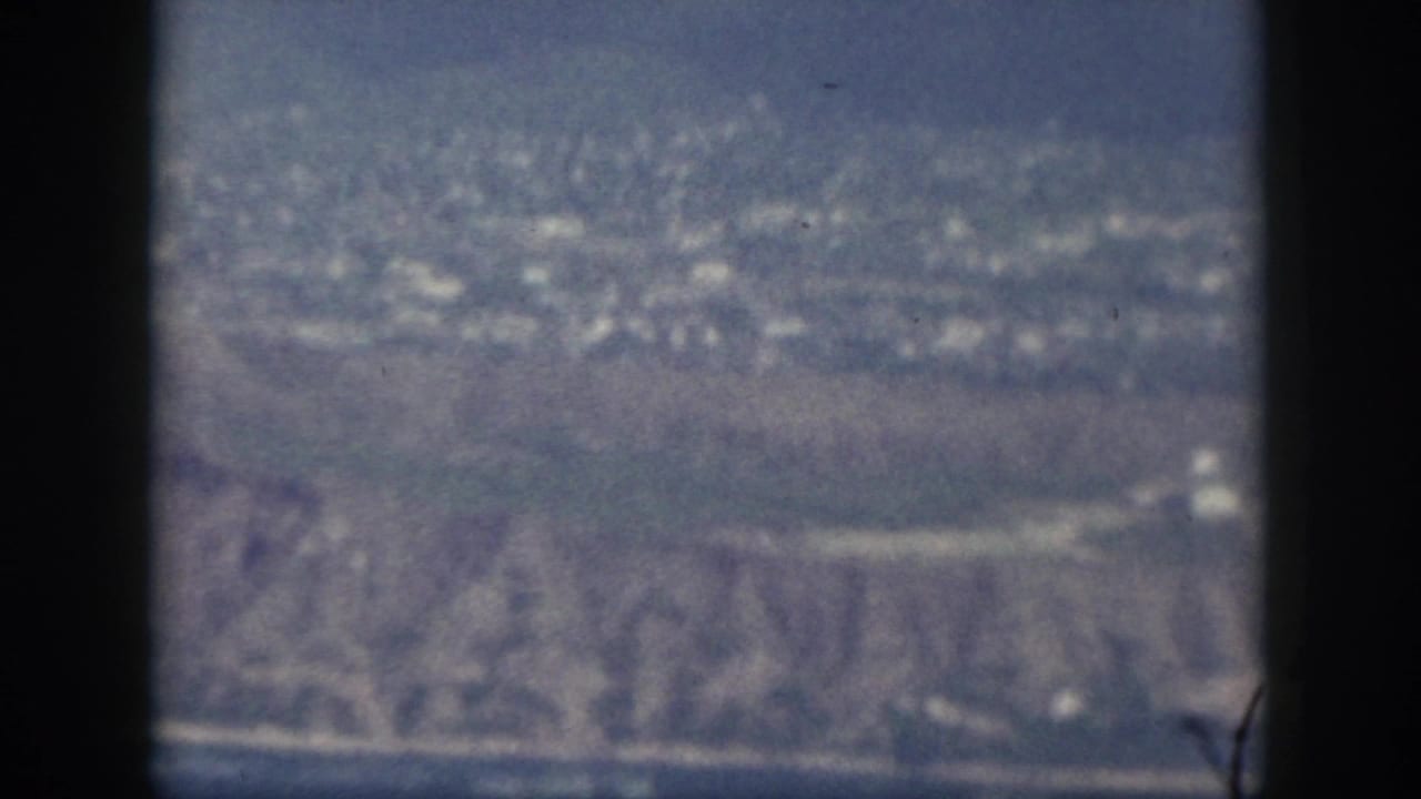 1959 Oahu aerial frame with dark window bars, blue sea, pale shoreline, and ridged coastal terrain in soft focus.