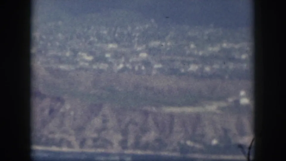 1959 Oahu aerial frame with dark window bars, blue sea, pale shoreline, and ridged coastal terrain in soft focus.