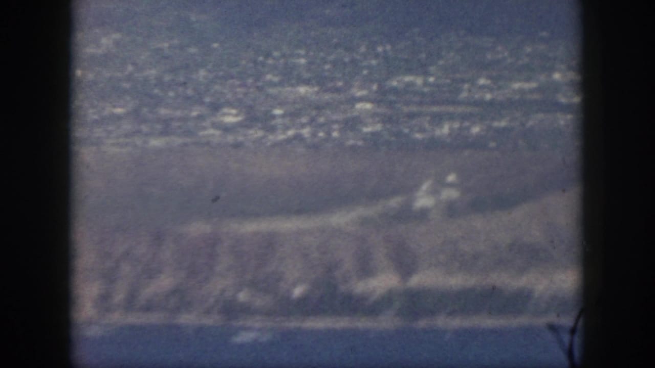1959 Oahu home-movie frame showing hazy coastal land, water, and distant development from a passenger airplane window.