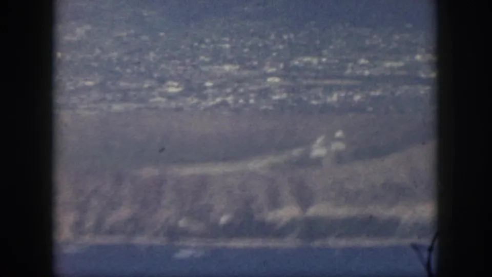 1959 Oahu home-movie frame showing hazy coastal land, water, and distant development from a passenger airplane window.