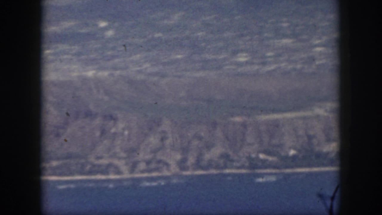 1959 Oahu aerial frame looking past airplane window edges toward water, shoreline, and hazy coastal terrain.