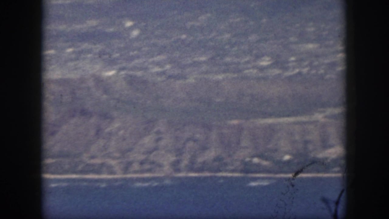1959 Oahu home-movie frame showing a hazy coastline beyond blue ocean, bordered by the dark film gate.