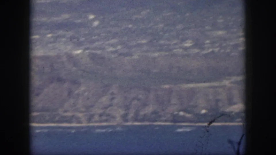 1959 Oahu home-movie frame showing a hazy coastline beyond blue ocean, bordered by the dark film gate.