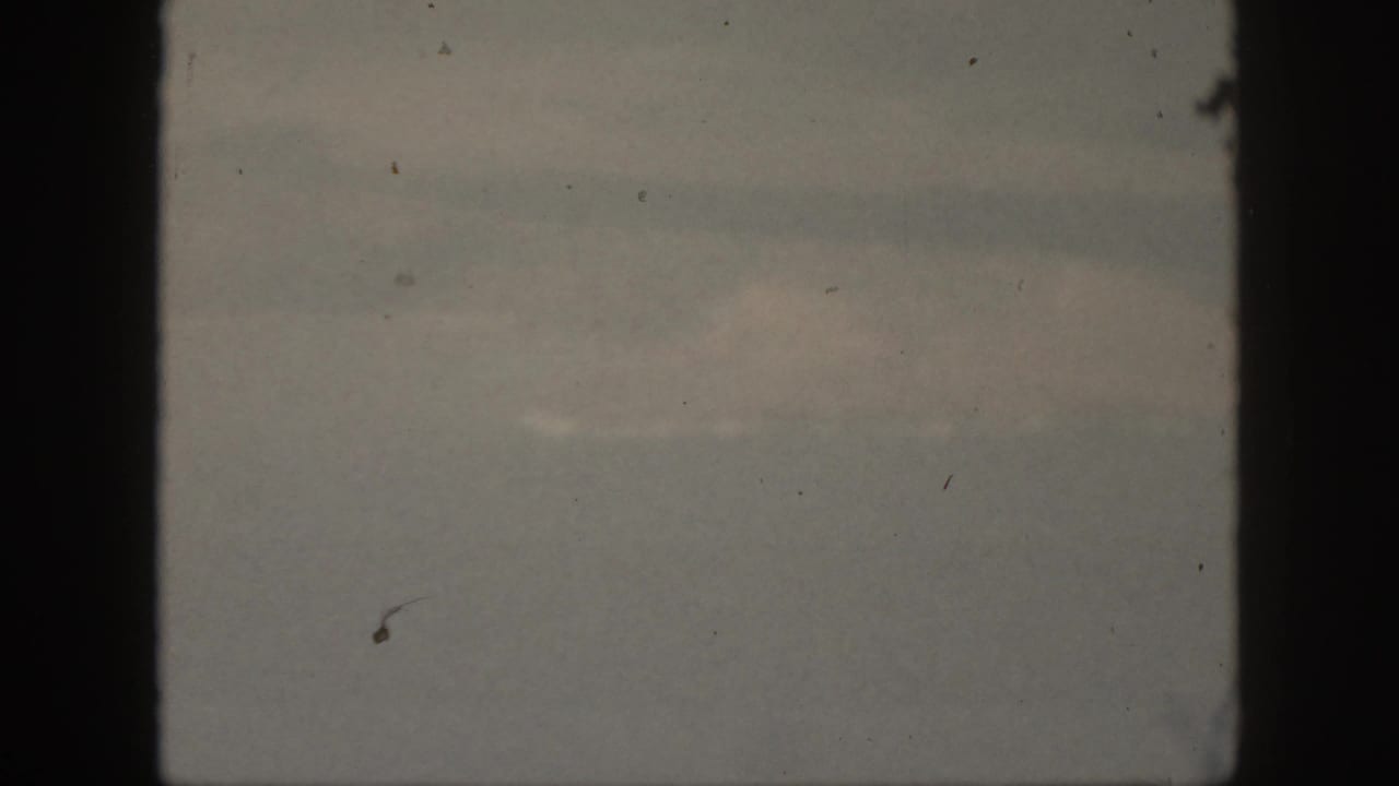 1959 Oahu 8mm frame showing a dim, overcast sky with soft cloud bands, black gate edges, visible dust, and muted color.