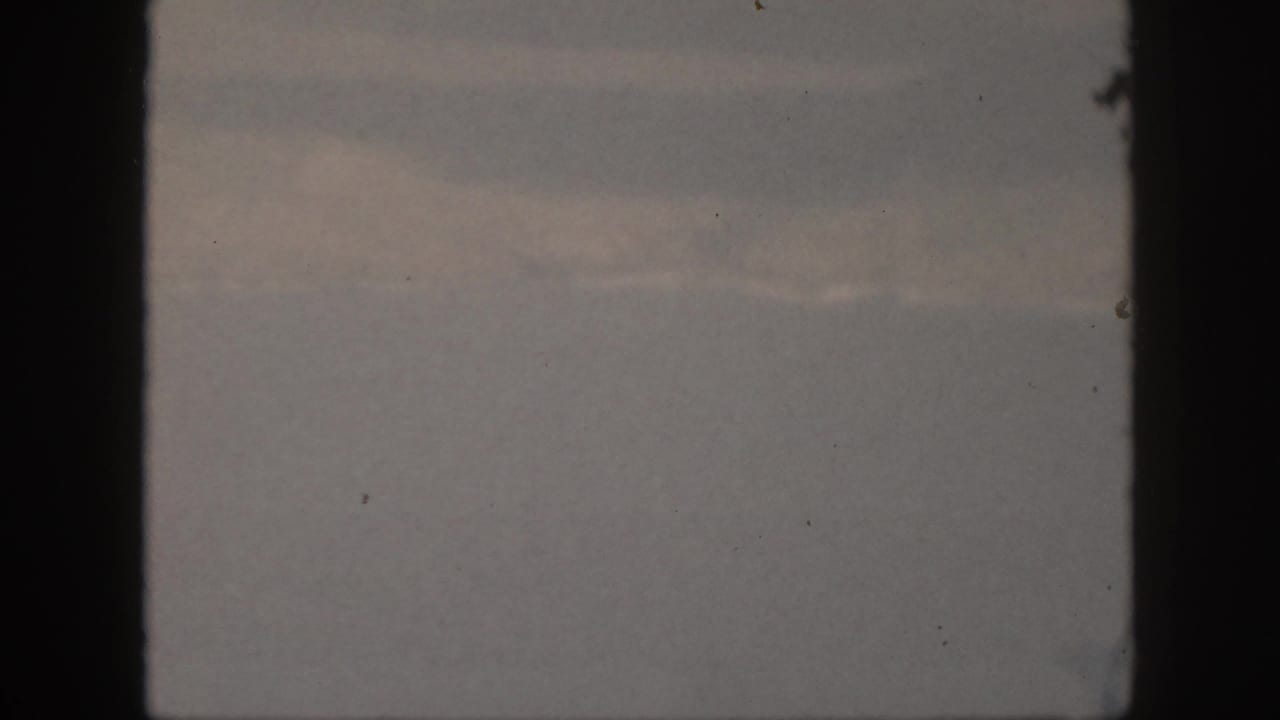 1959 Oahu 8mm still showing blue-gray cloud cover, subdued light patches, black frame edges, grain, and small dust marks.