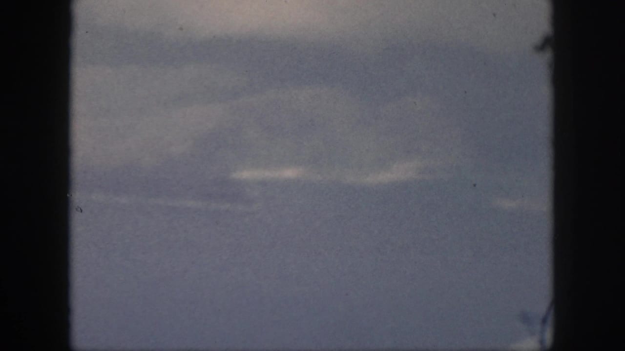 1959 Oahu 8mm frame showing layered dark clouds with small bright breaks, blue-gray tones, dust specks, and heavy black gate borders.