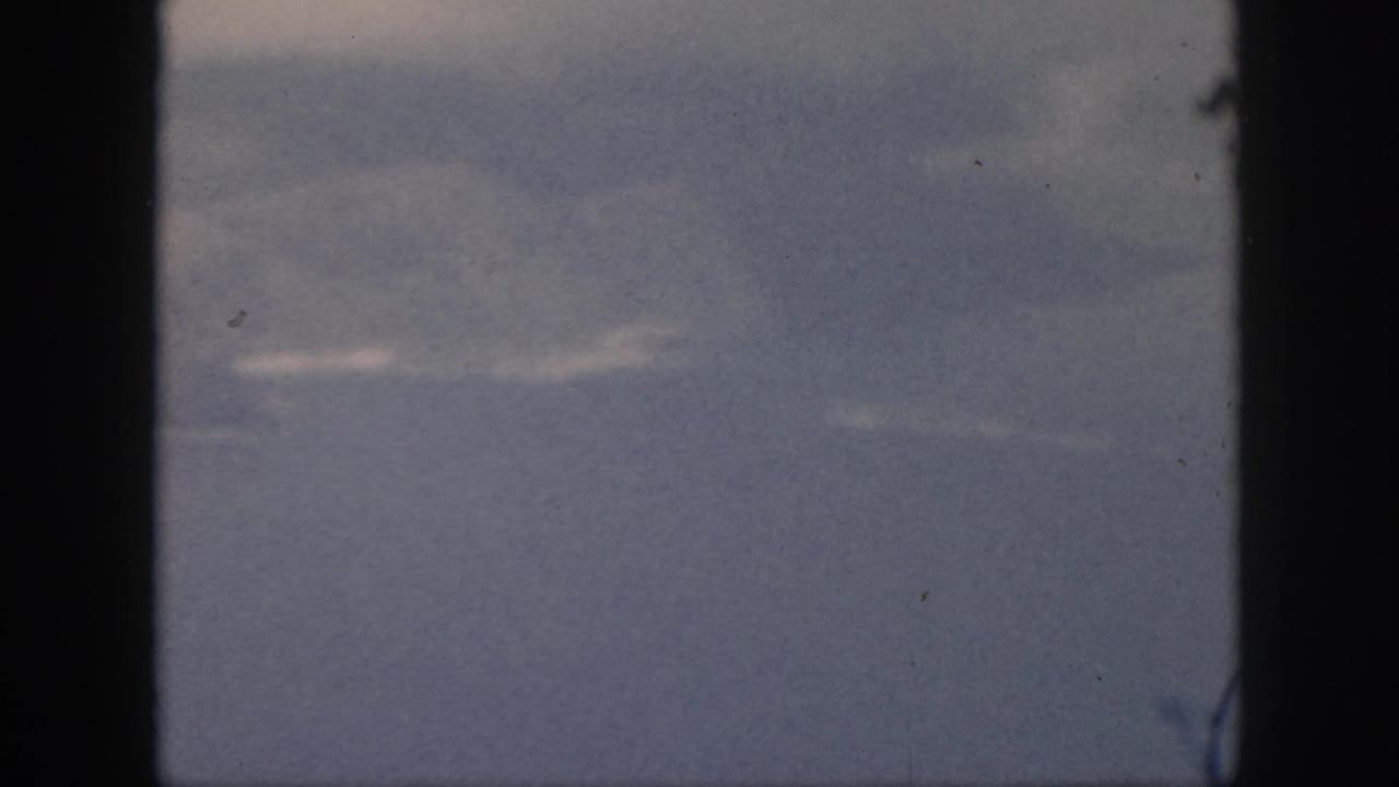1959 Oahu home-movie frame looking through an airplane window toward blue-gray sea or sky and thin cloud streaks.