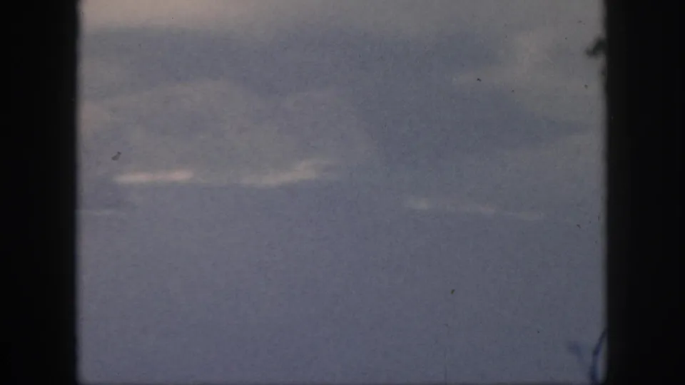 1959 Oahu home-movie frame looking through an airplane window toward blue-gray sea or sky and thin cloud streaks.
