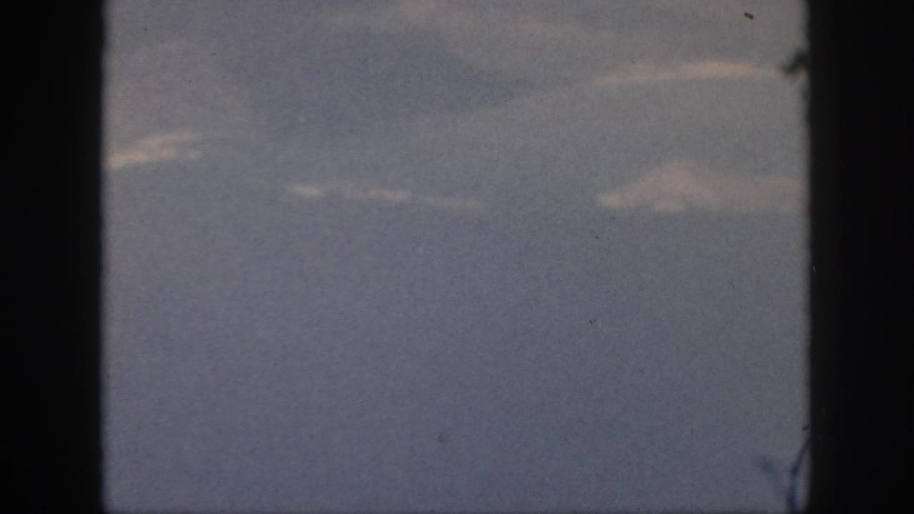 1959 Oahu travel footage frame showing a muted ocean-and-sky view through dark aircraft window framing.