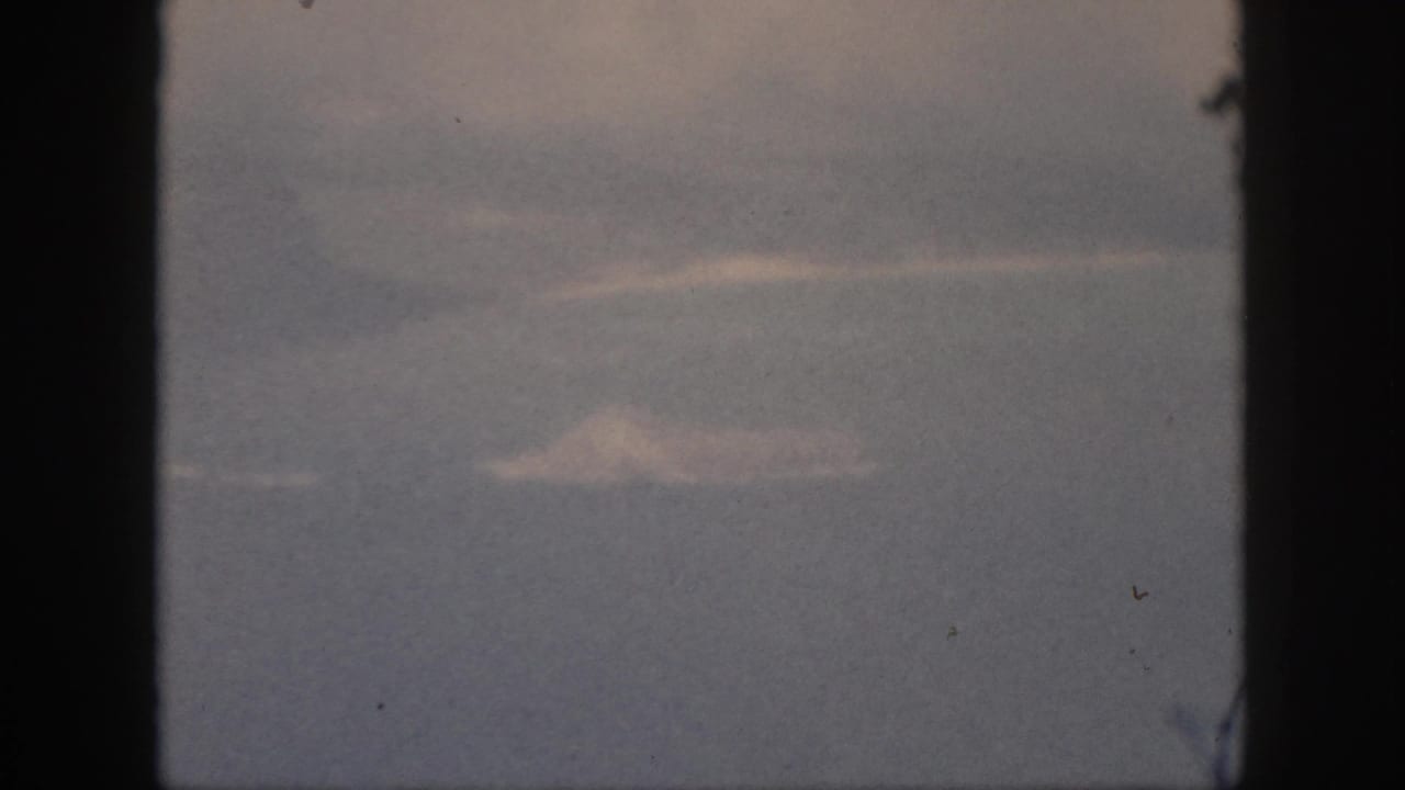 Aircraft-window view from 1959 Hawaii travel footage, with pale cloud shapes and a dark window edge dominating the frame.