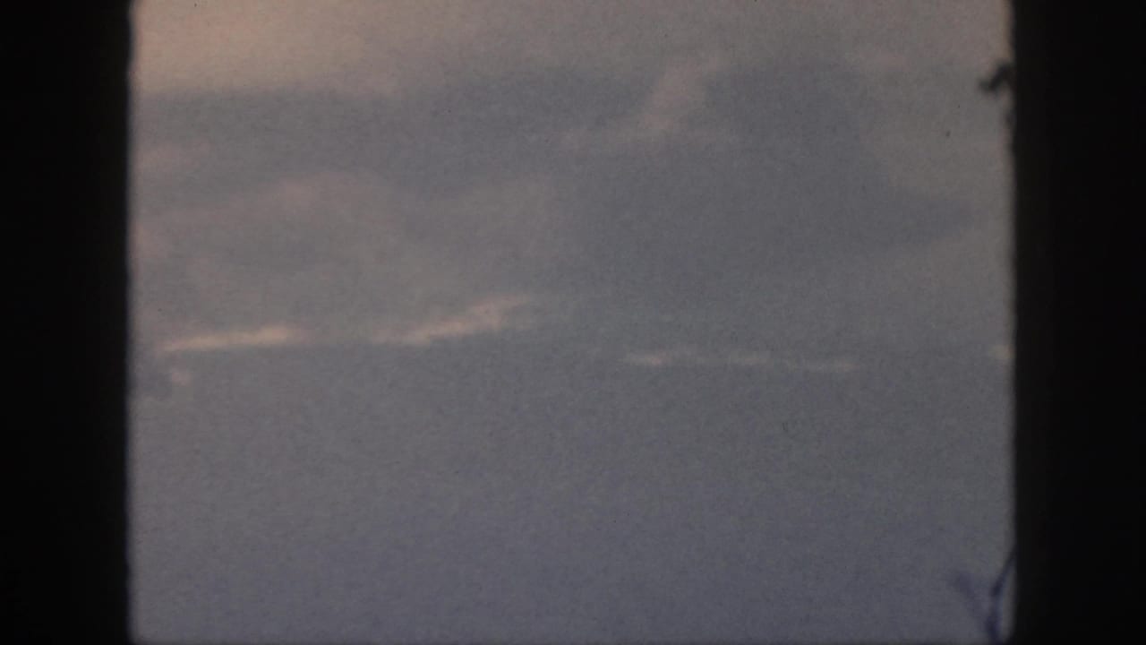8mm airplane passenger view with blue-gray haze, faint cloud bands, and heavy dark window framing.