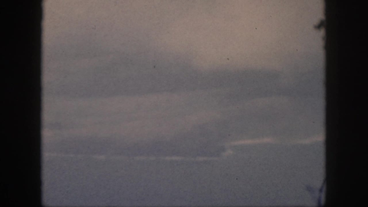 1959 Oahu home-movie frame showing a soft, hazy aerial view through dark window framing, with faint clouds and muted blue-gray distance.