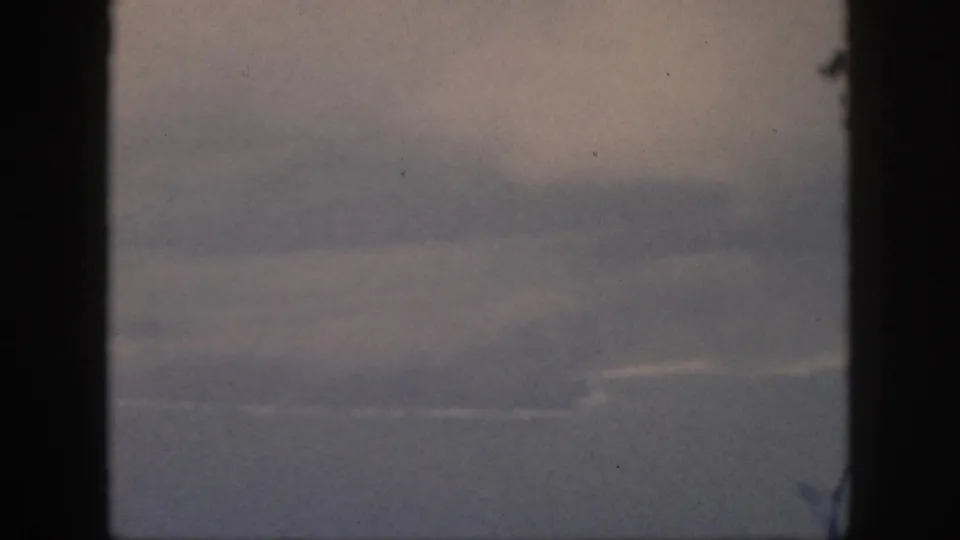 1959 Oahu home-movie frame showing a soft, hazy aerial view through dark window framing, with faint clouds and muted blue-gray distance.