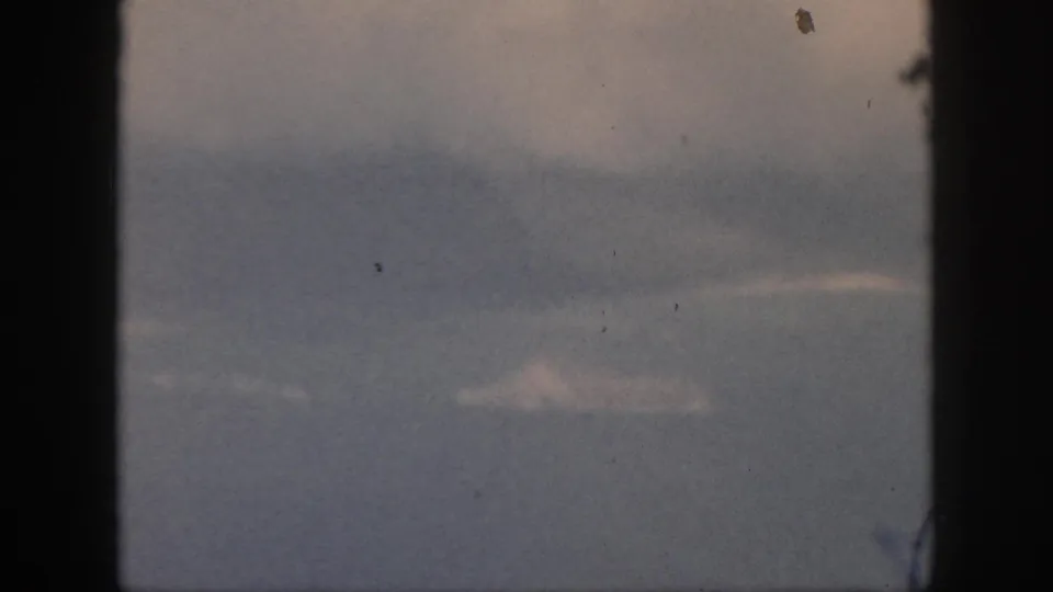 Oahu aerial footage from 1959 appears through dark window edges, with pale clouds and an indistinct blue-gray landscape or sea below.