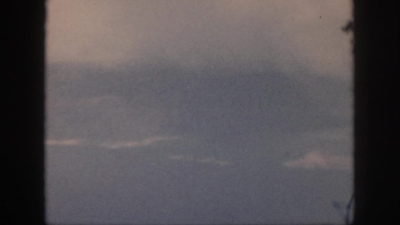 A faded 8mm frame from Oahu shows a hazy aerial view through window bars, with faint cloud bands and indistinct terrain or water.