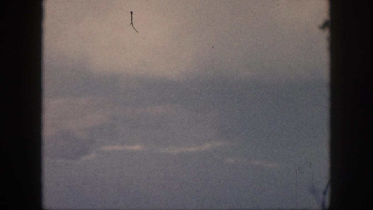 The final 1959 Oahu frame holds on a hazy aerial scene through a dark aircraft window, with pale cloud streaks and visible 8mm dust.