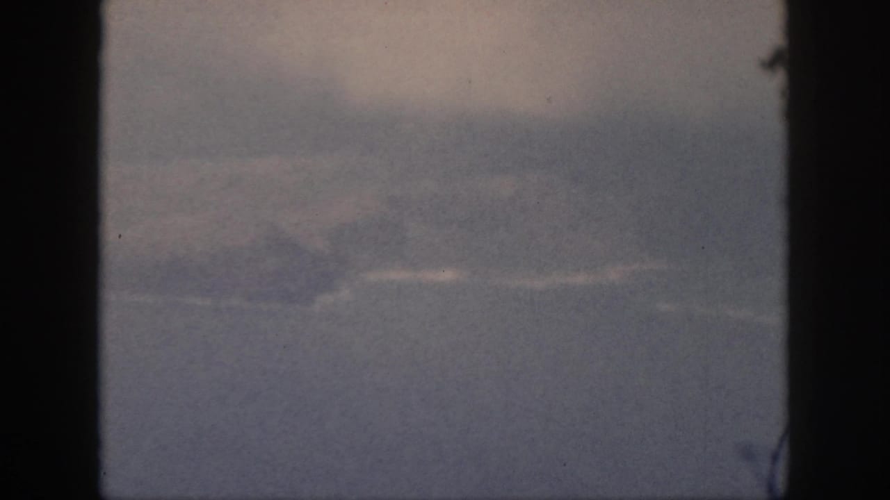 1959 Oahu home-movie frame showing a hazy ocean horizon and faint landform through dark vertical window edges.