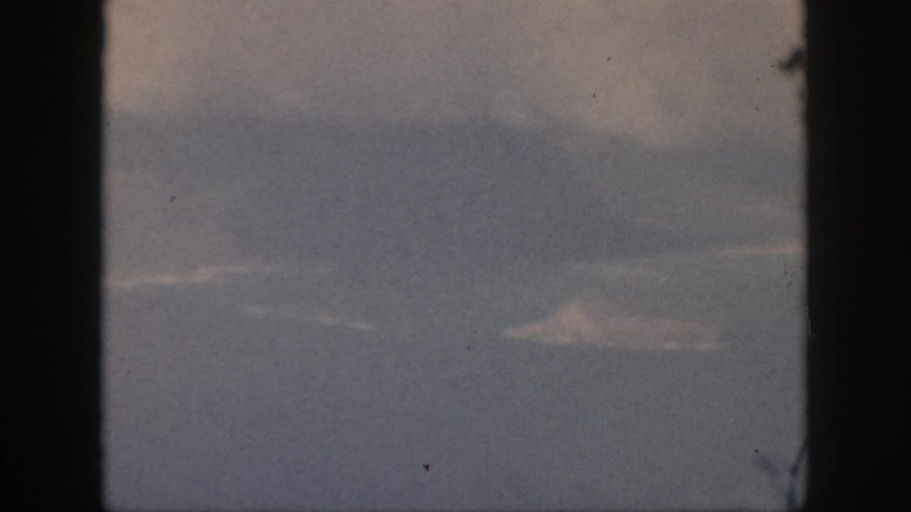 1959 Oahu travel footage frame with a pale distant landform, cloud cover, and dark window framing at the sides.
