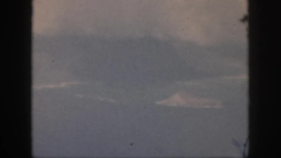 1959 Oahu travel footage frame with a pale distant landform, cloud cover, and dark window framing at the sides.
