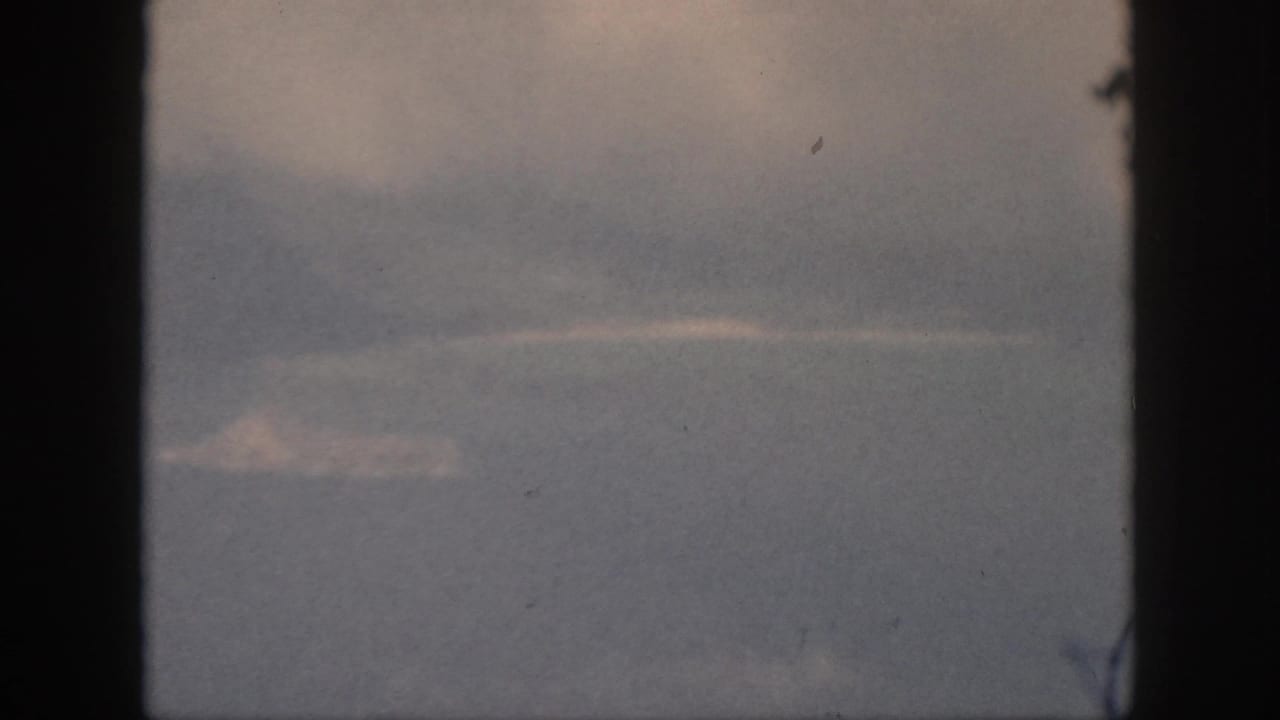 1959 Oahu 8mm frame showing a small pale landform beneath low clouds, viewed through a window with faded color.