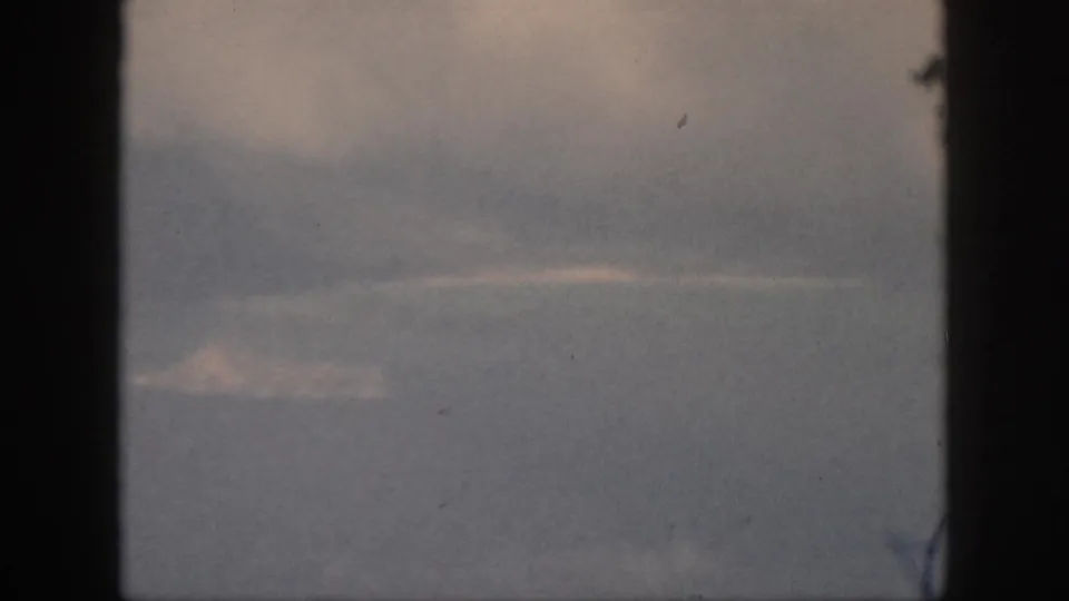 1959 Oahu 8mm frame showing a small pale landform beneath low clouds, viewed through a window with faded color.