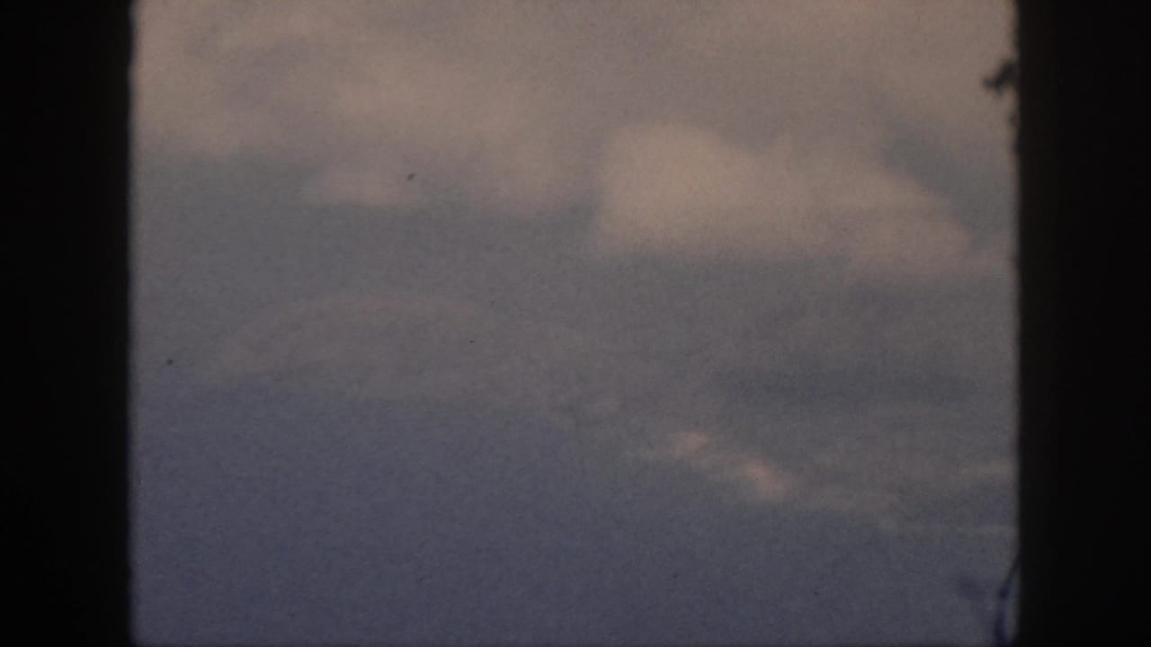 1959 Oahu 8mm frame showing hazy sky, soft clouds, and a faint distant coast or water line bordered by dark film edges.
