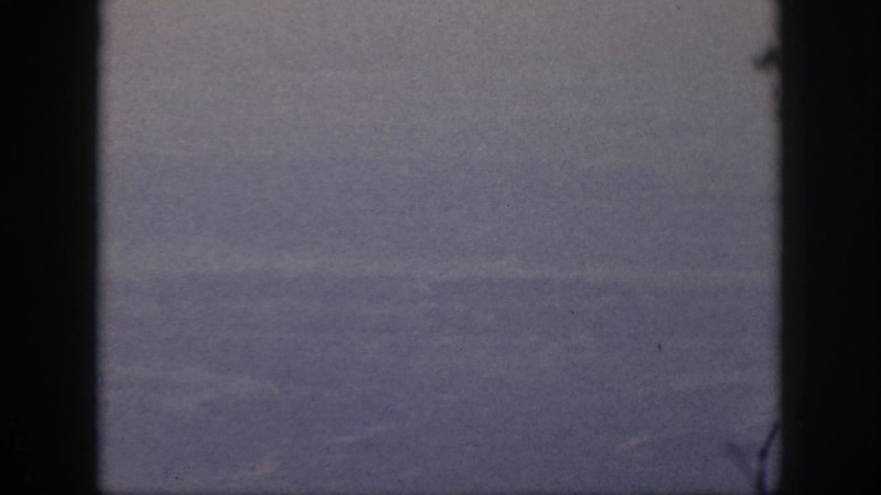 1959 Oahu flight view showing indistinct ground below through a dark airplane window frame on grainy 8mm film.