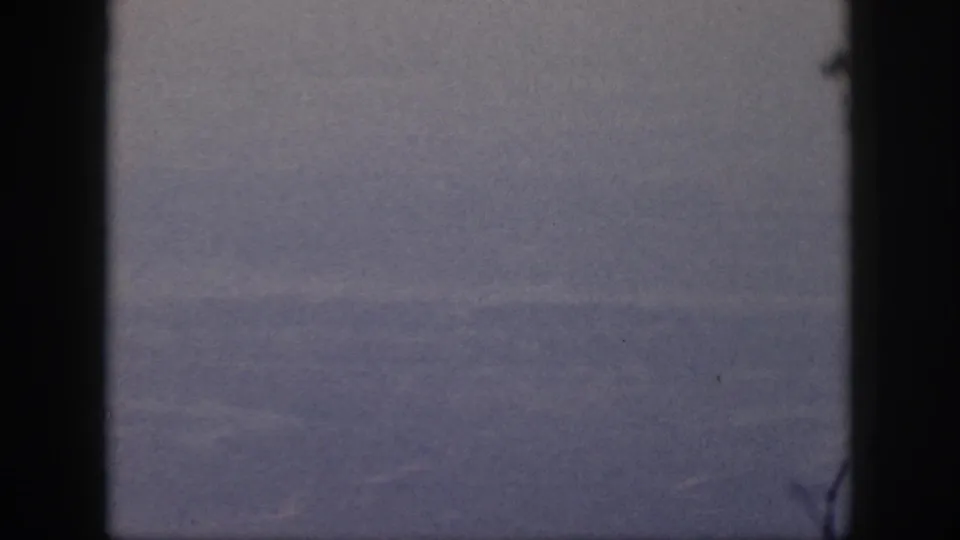 1959 Oahu flight view showing indistinct ground below through a dark airplane window frame on grainy 8mm film.