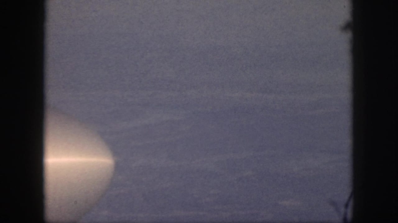 1959 Oahu home-movie frame from an airplane window, showing hazy sky and a pale aircraft form at the edge.