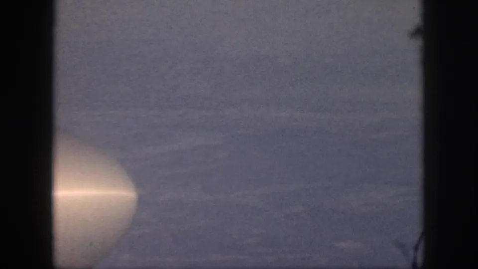1959 Oahu home-movie frame from an airplane window, showing hazy sky and a pale aircraft form at the edge.