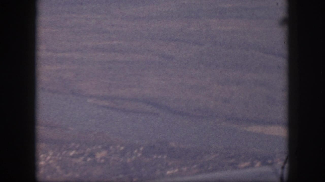 1959 Oahu aerial home-movie view showing coastline, fields or terrain, and a distant town beyond the aircraft window frame.