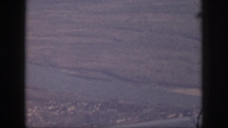 1959 Oahu aerial home-movie view showing coastline, fields or terrain, and a distant town beyond the aircraft window frame.