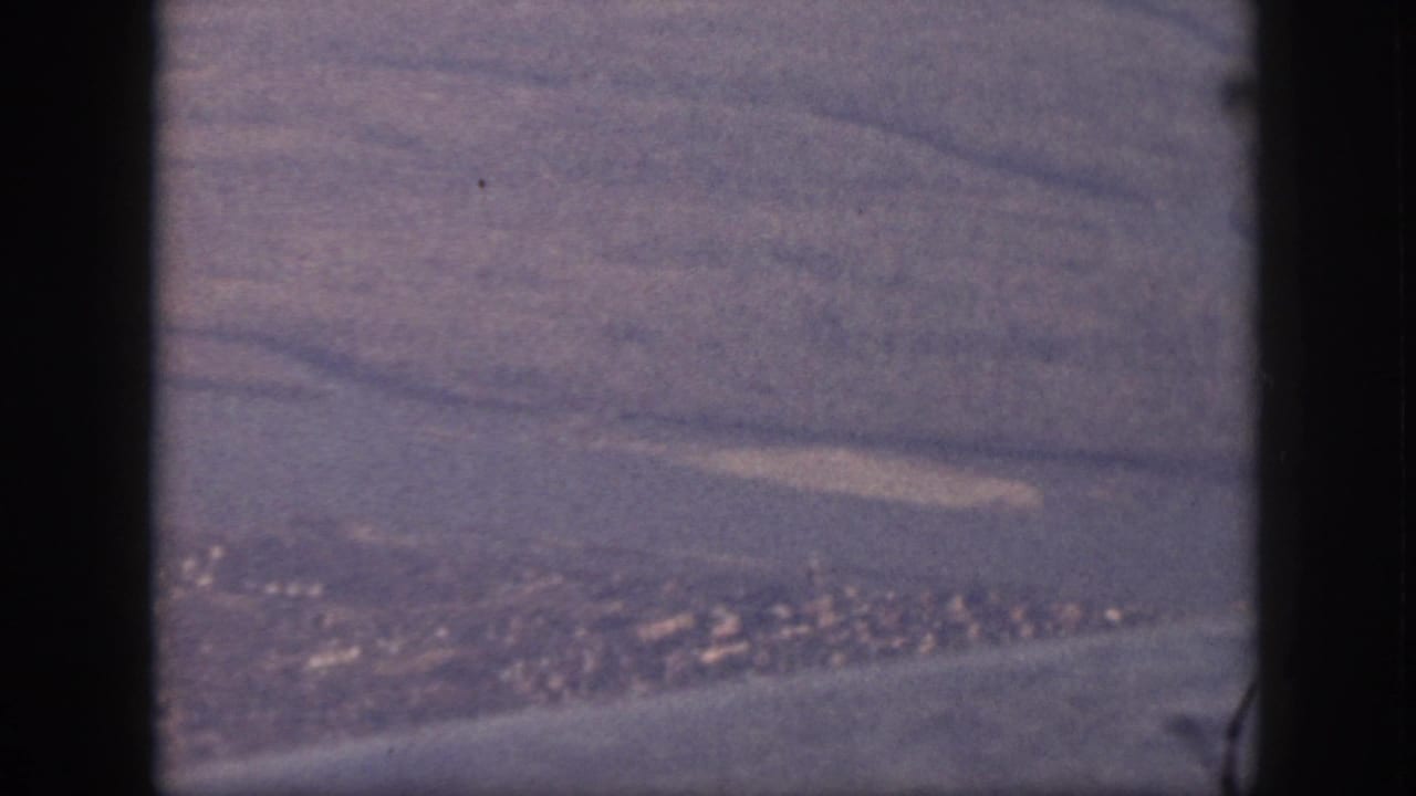 Oahu aerial frame from 1959 showing coastal terrain, water, and a distant town or village through an airplane window.