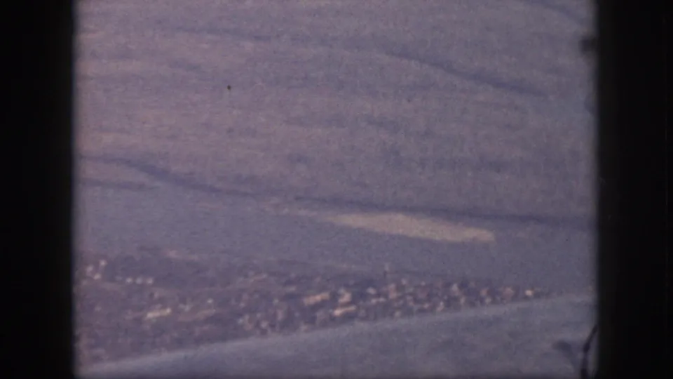 Oahu aerial frame from 1959 showing coastal terrain, water, and a distant town or village through an airplane window.