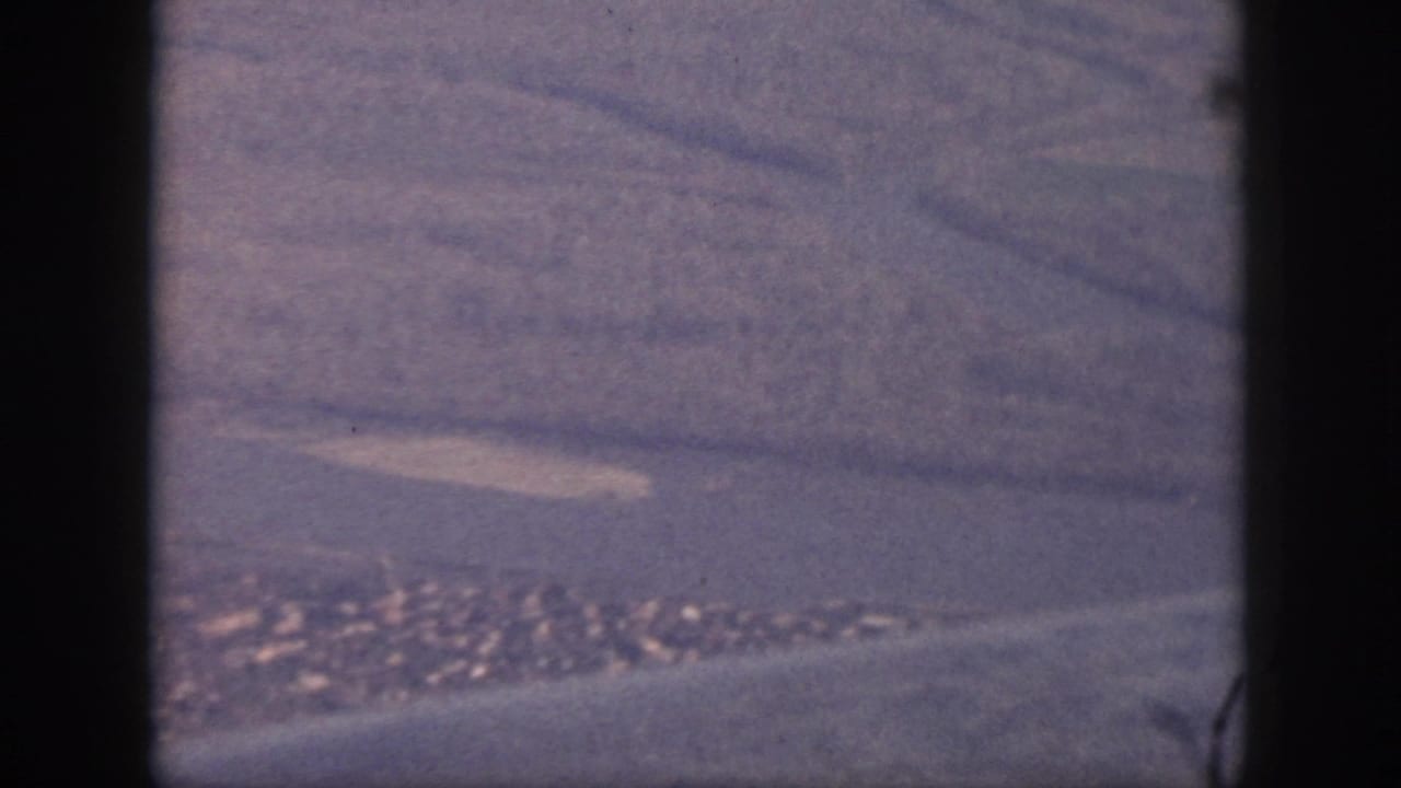 Aerial 8mm view over Oahu, with a pale shoreline, small developed area, water, and dark aircraft-window or gate-edge framing.